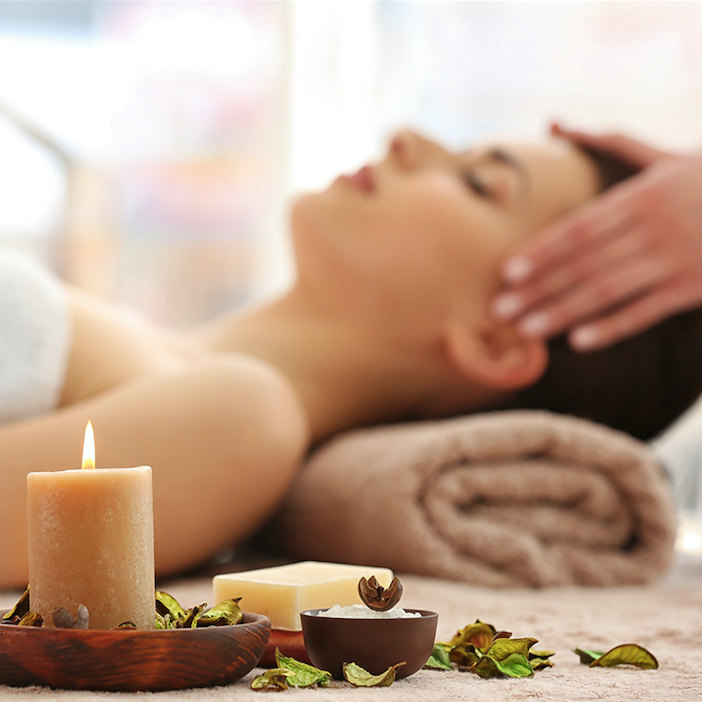 Woman lying on a spa table receiving a head massage with a lit candle and bowls of aromatherapy ingredients in the foreground