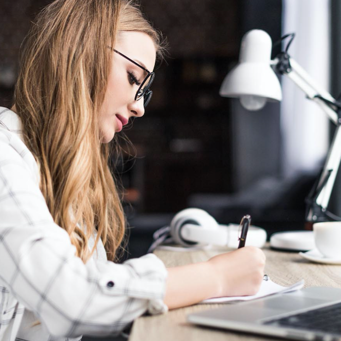 Woman writing notes at a desk with laptop, headphones, and desk lamp in a focused study setting