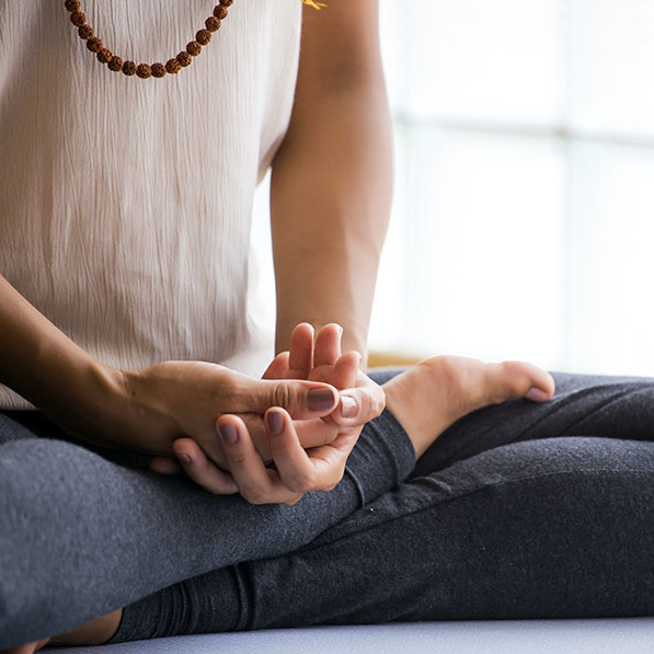 Person sitting cross-legged meditating with hands in a mudra and mala beads by a sunlit window