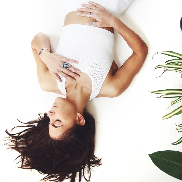 Woman lying on her back with hand on chest practicing deep breathing on a white floor beside a potted plant