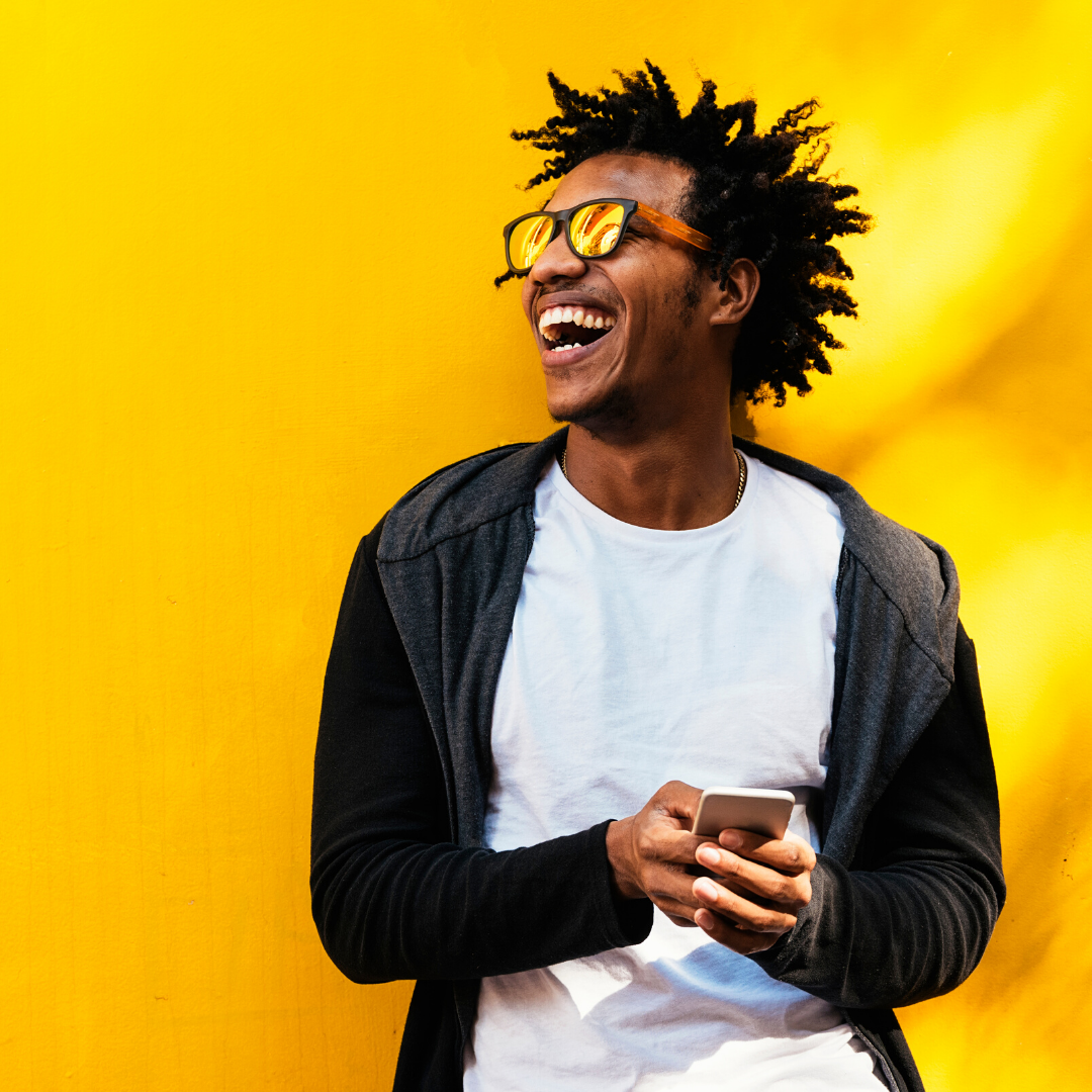 Young man laughing while holding a smartphone against a bright yellow wall