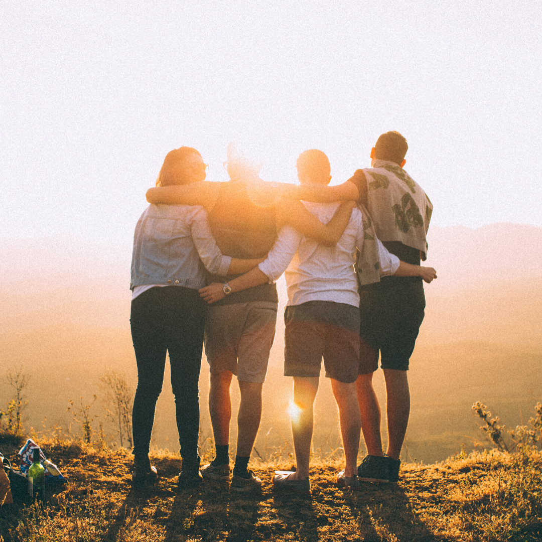 Four friends standing arm-in-arm on a grassy hilltop watching the sun set over a valley