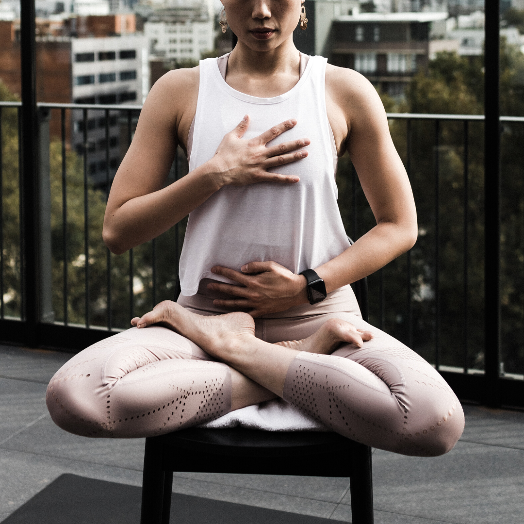Woman seated cross-legged on a stool practicing breathwork with hands on chest and belly on a rooftop balcony with city buildings behind