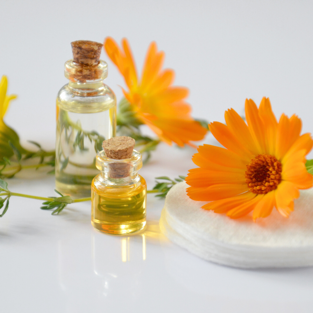 Two small glass bottles of essential oils arranged with orange calendula flowers and cotton pads on a white surface