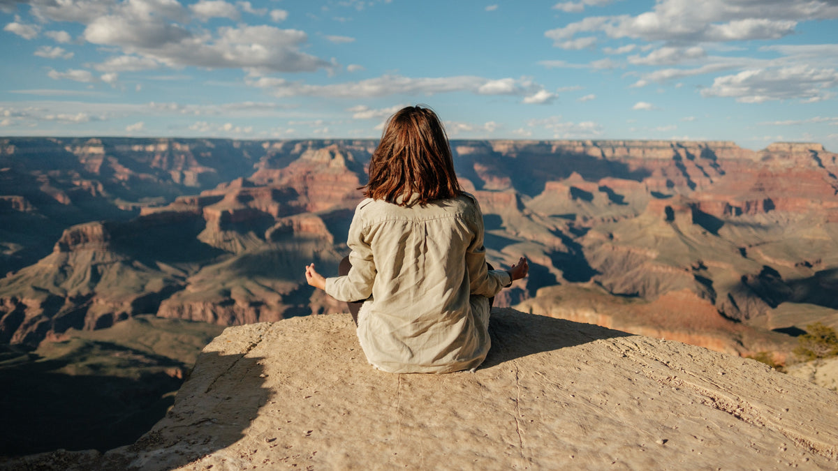 Woman meditating cross-legged on a rocky canyon rim overlooking a sunlit expansive canyon and blue sky