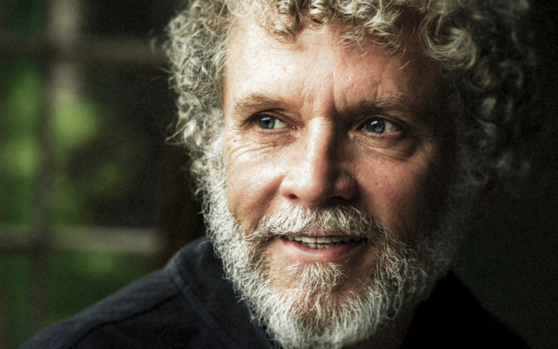Older man with curly gray hair and beard looking thoughtfully off to the side in a softly lit indoor setting