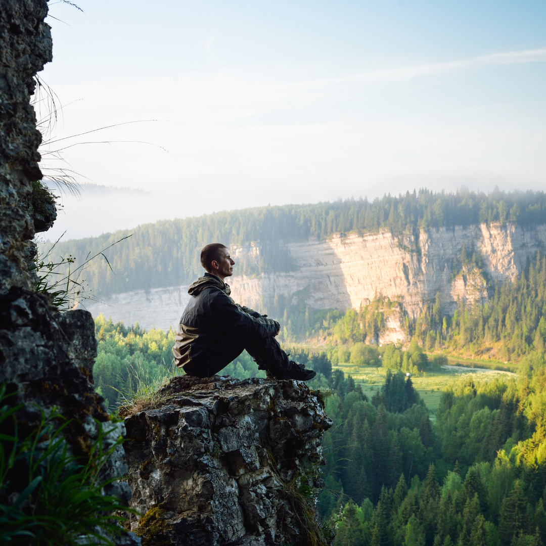 Person sitting on a rocky cliff edge overlooking a sunlit forested valley, quietly breathing and reflecting
