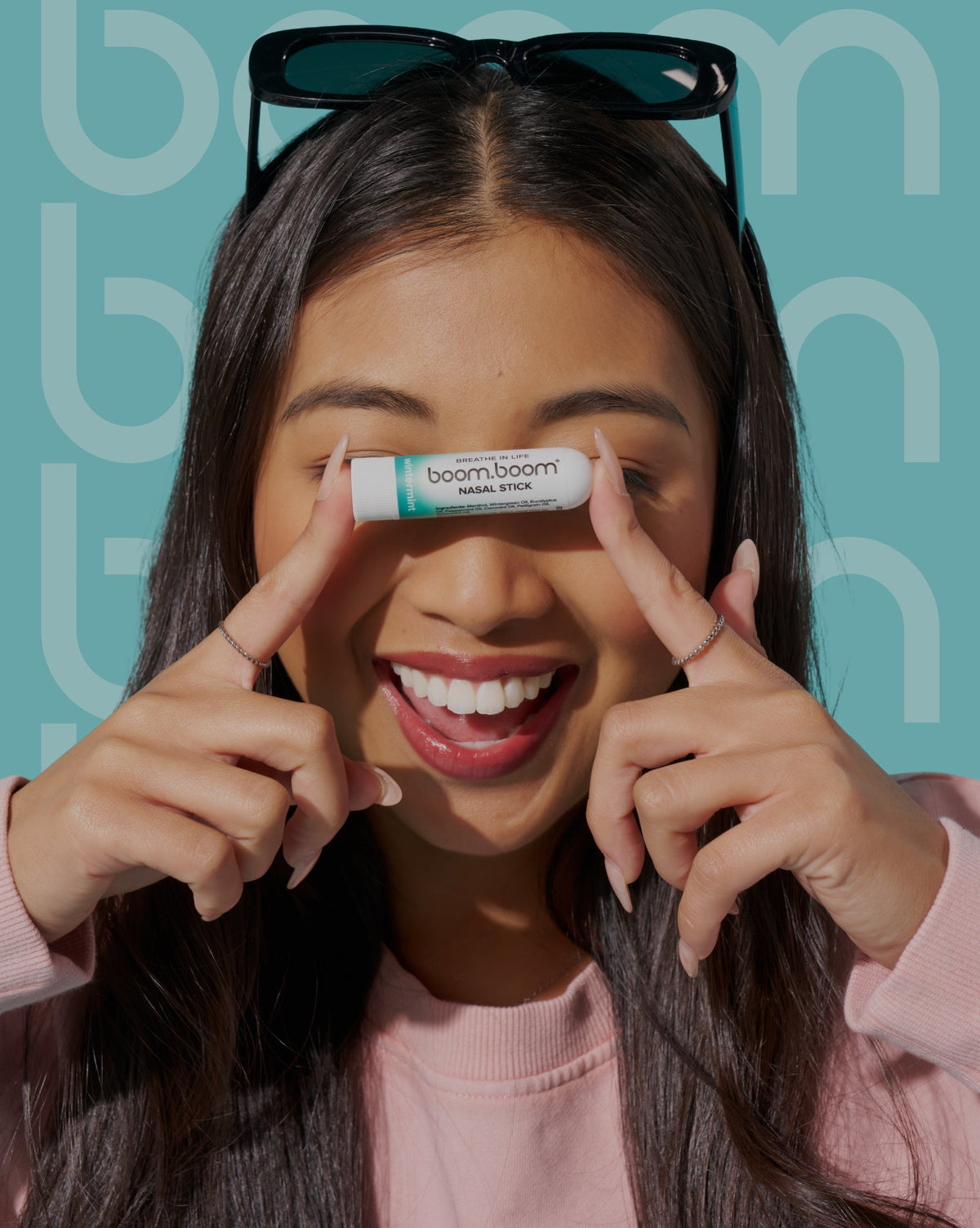 Young woman playfully holding a BoomBoom nasal stick under her nose while smiling against a teal branded background