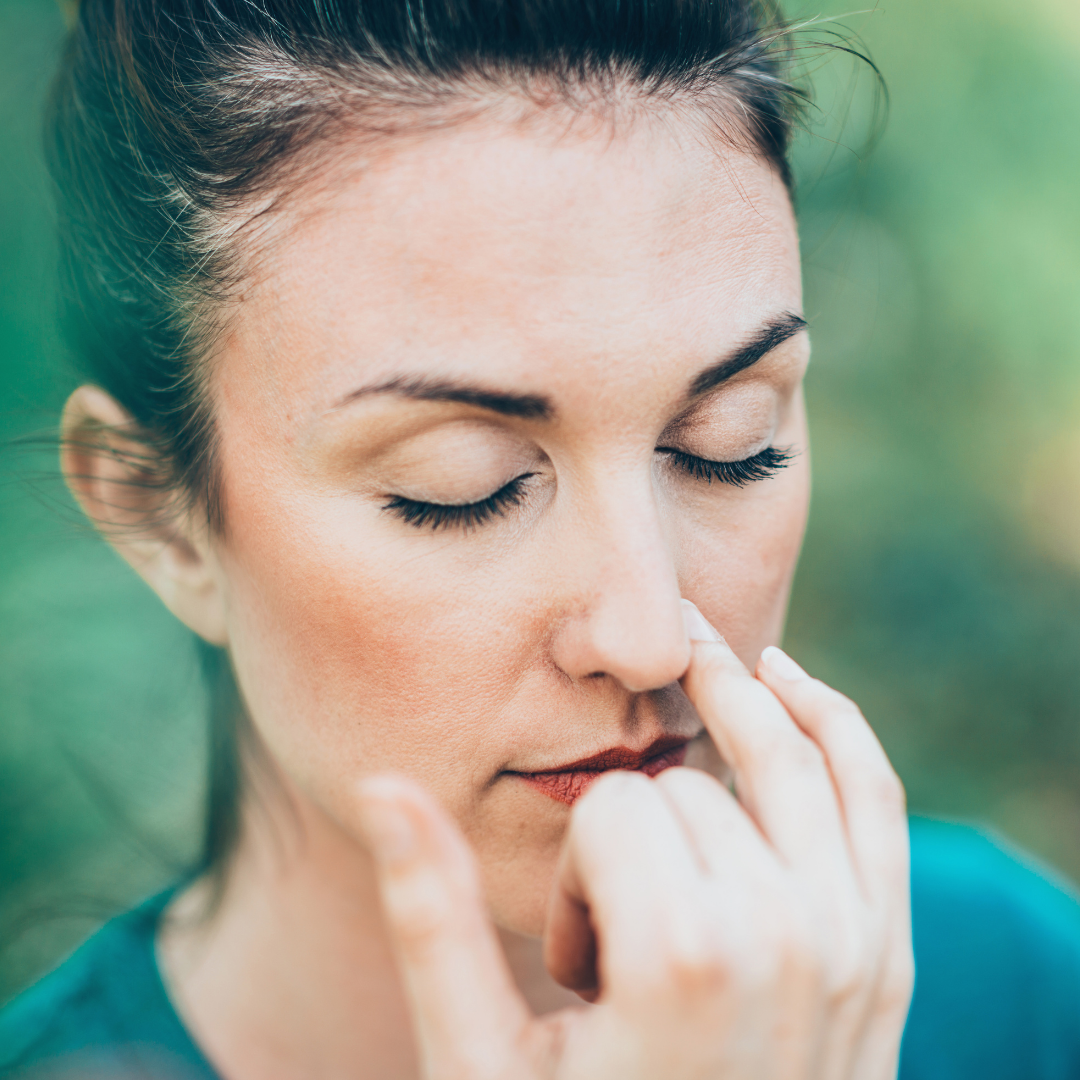 Woman pinching one nostril and inhaling through the other with eyes closed against a blurred green background