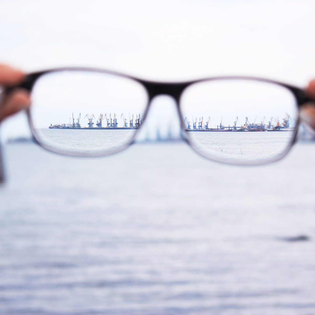 Eyeglasses held up to show distant harbor cranes in sharp focus against a blurred sea background