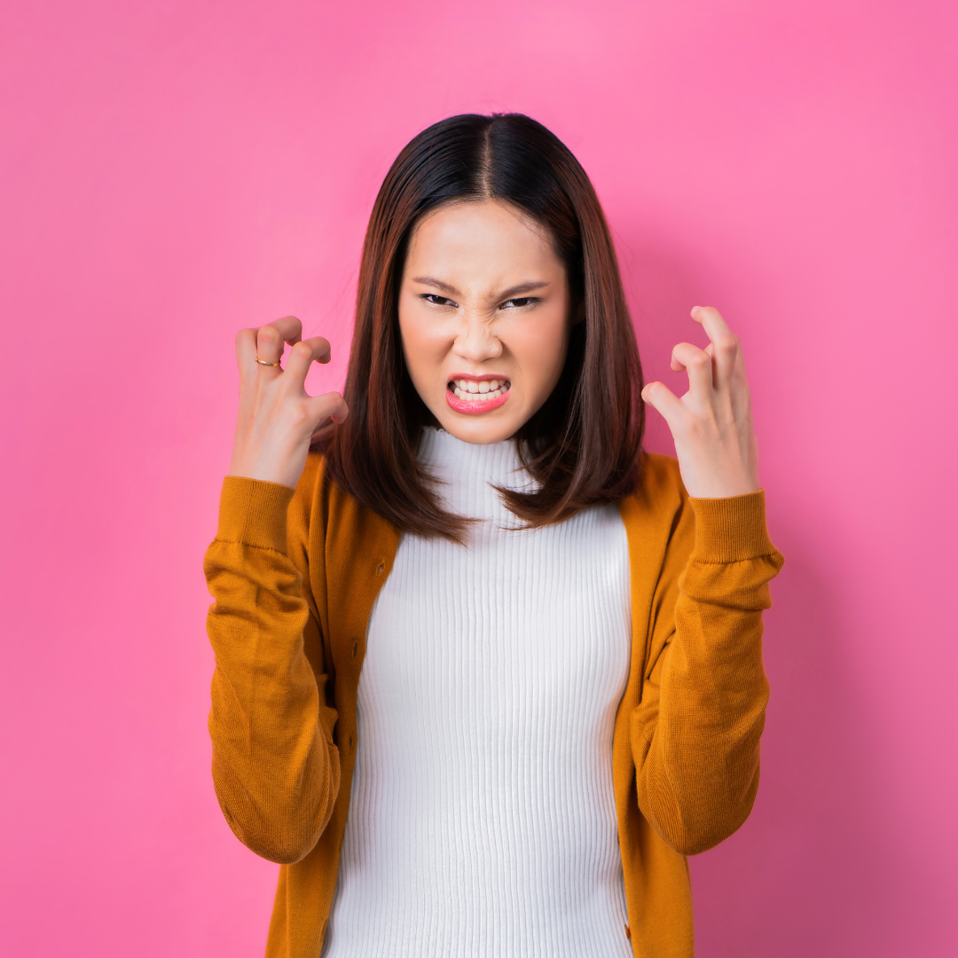 Young woman in a mustard cardigan baring her teeth and making clawing gestures against a solid pink background