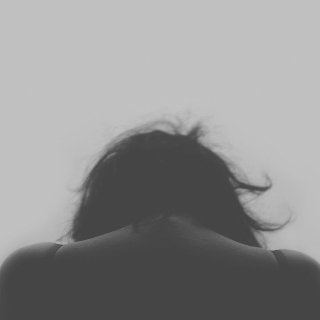 Person seated with head bowed facing away, shoulders visible against a muted gray background conveying grief and solitude