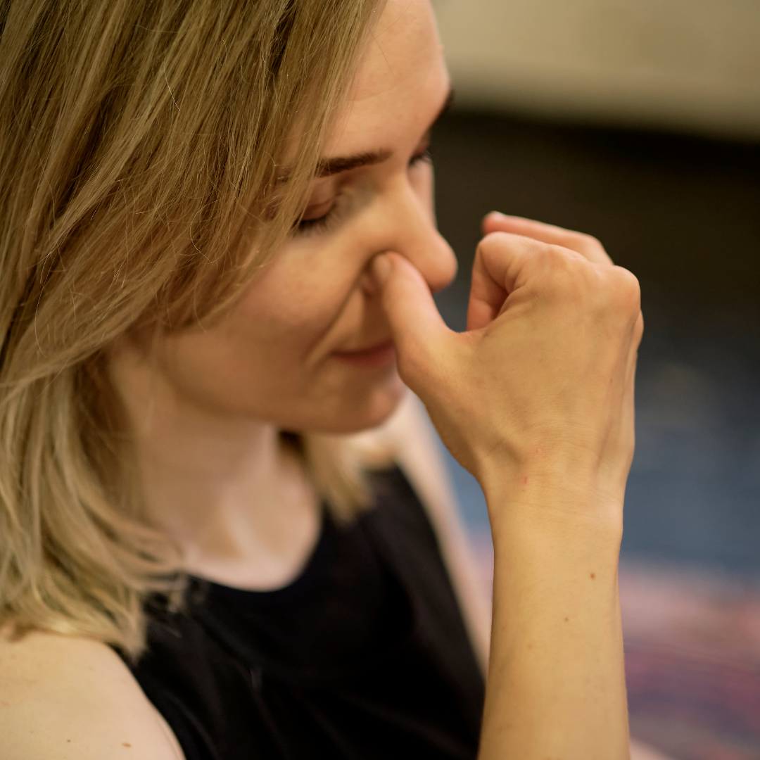 Woman pinching one nostril while practicing a nose-breathing exercise, seated indoors with eyes closed