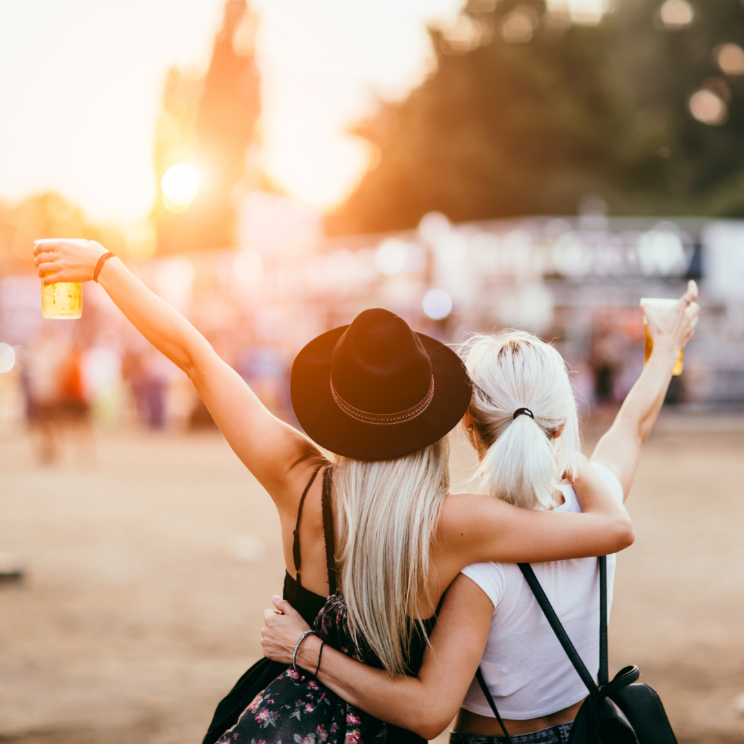 Two friends with arms around each other raising drinks at an outdoor music festival at sunset