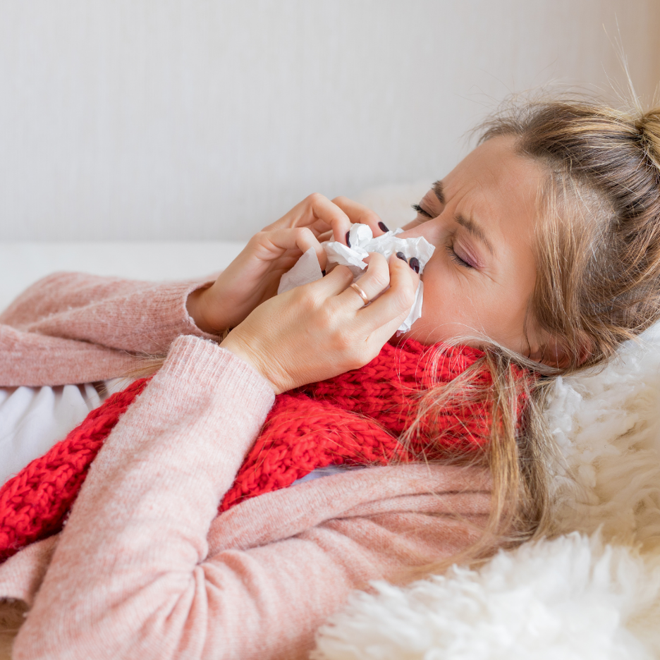 Woman lying in bed blowing her nose into a tissue, wrapped in a red scarf and pink sweater