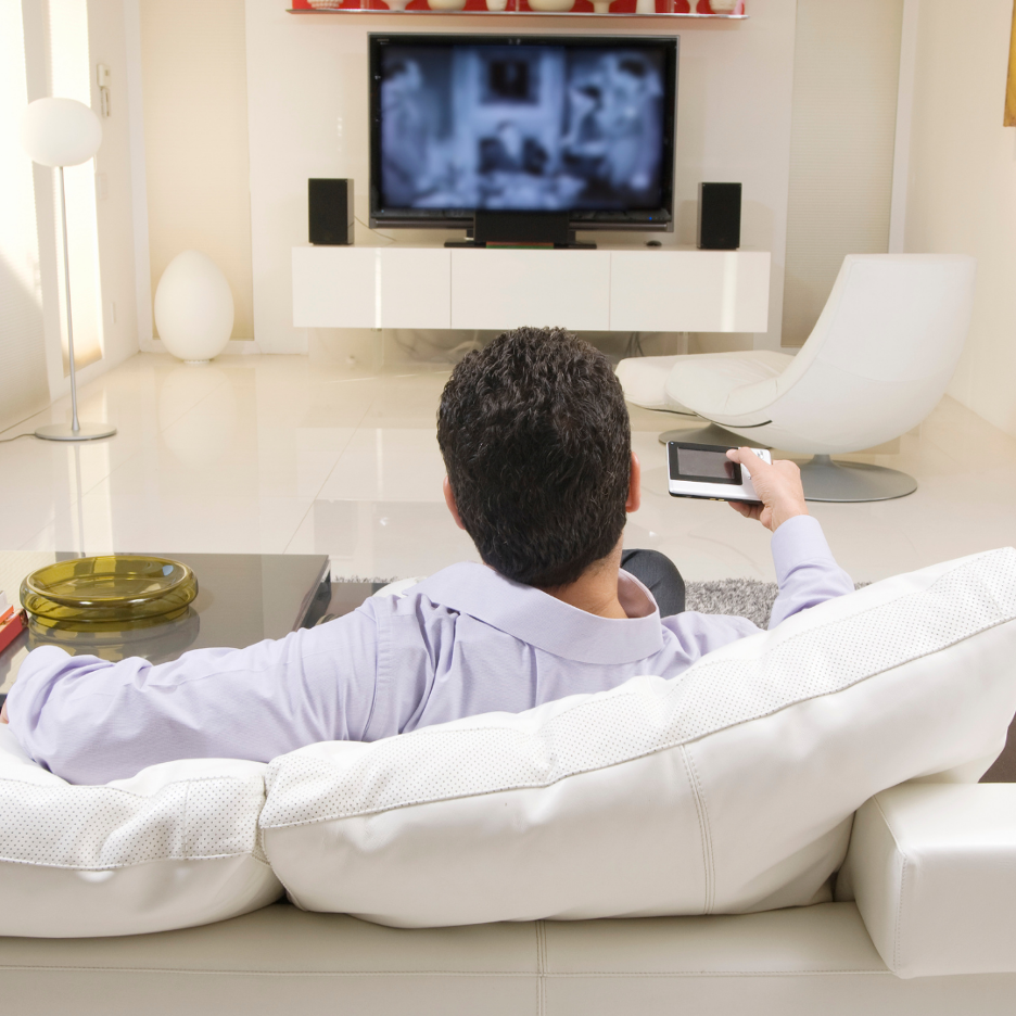 Man sitting on a white couch holding a remote and watching TV in a bright modern living room