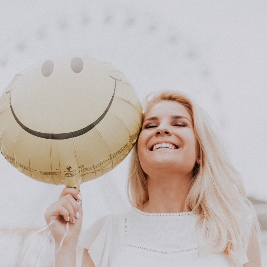 Young woman smiling while holding a yellow smiley-face balloon against a pale outdoor background