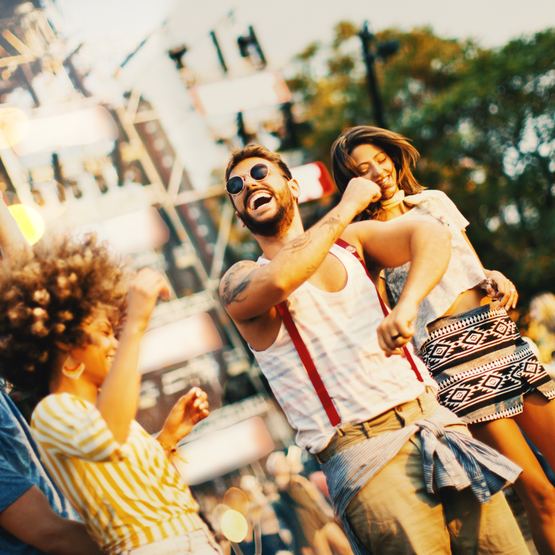 Young people dancing and laughing near a lit outdoor concert stage at a music festival