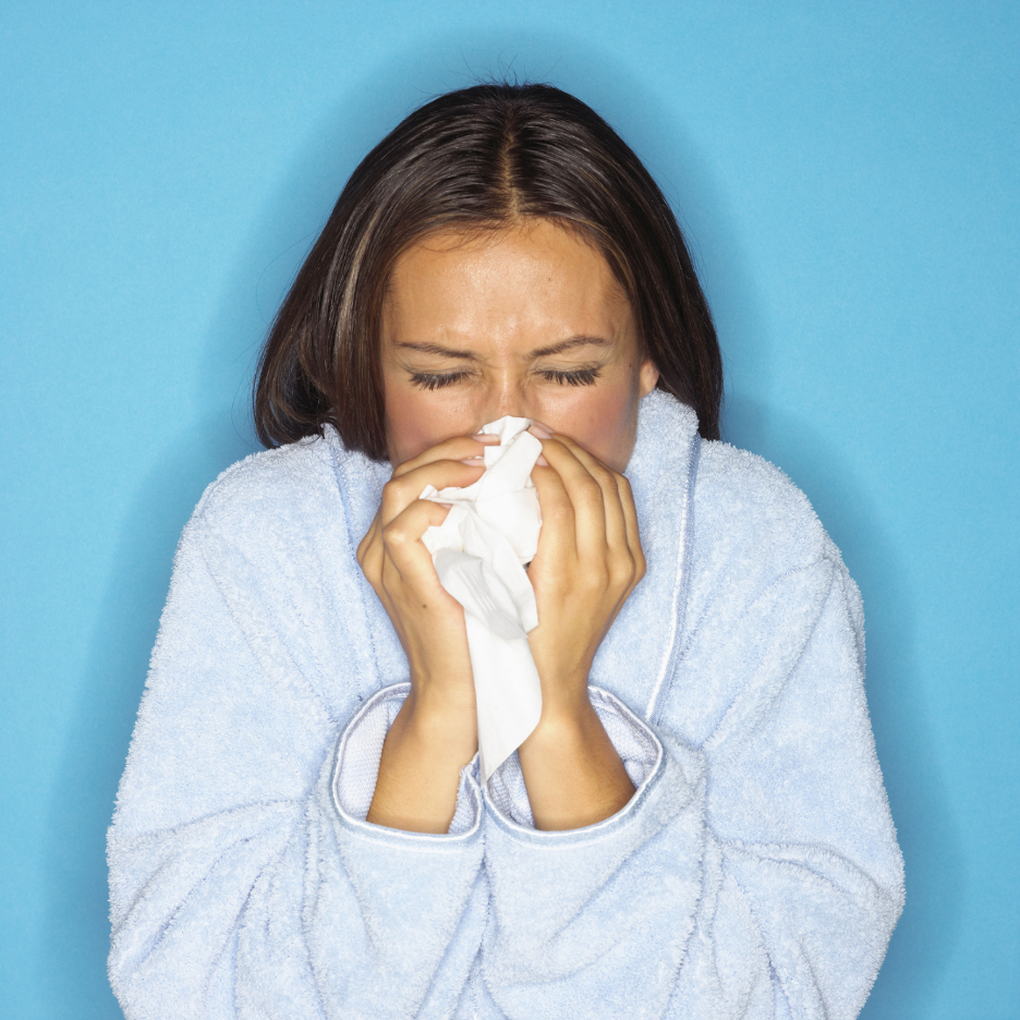 Woman sneezing into a tissue while wearing a light blue robe against a blue background