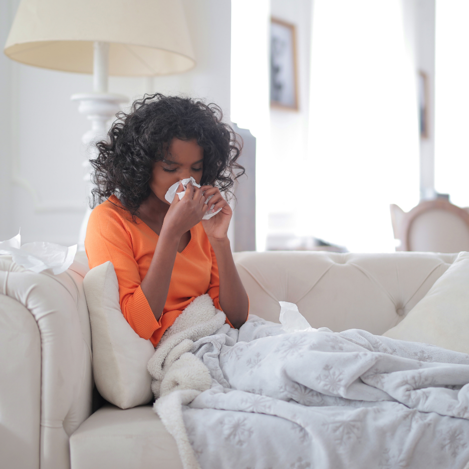 Woman sitting on a couch blowing her nose into a tissue, wrapped in a blanket in a bright living room