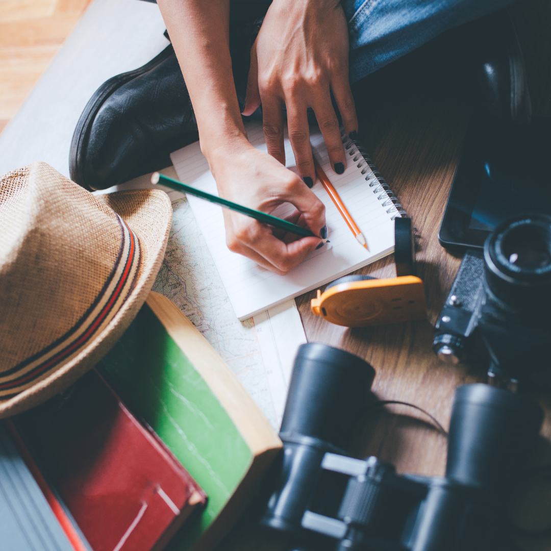 Hands writing a packing list in a notebook surrounded by a straw hat, map, camera, binoculars, and books on a wooden floor
