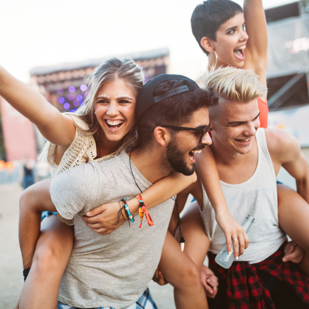 Four friends laughing, two giving piggyback rides at an outdoor music festival with a blurred stage in the background