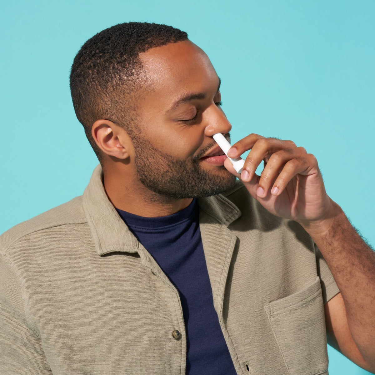 Man holding a white, pocket-sized nasal stick to his nostril with eyes closed against a turquoise background