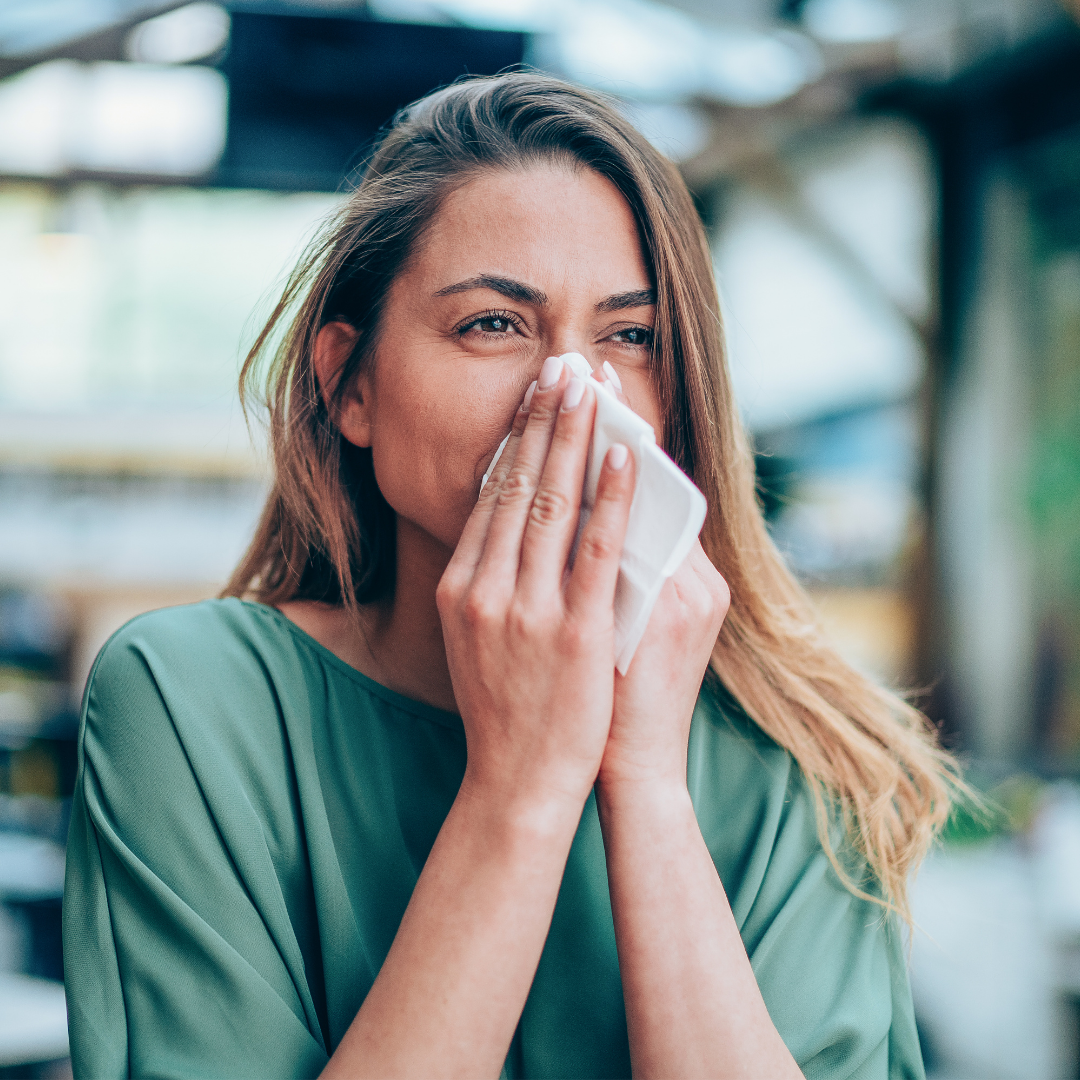 Woman blowing her nose into a tissue, showing allergy symptoms in a bright indoor setting