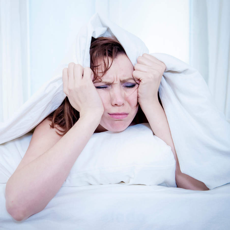 Woman in bed pulling blanket over her head, wincing and appearing congested from nighttime allergies