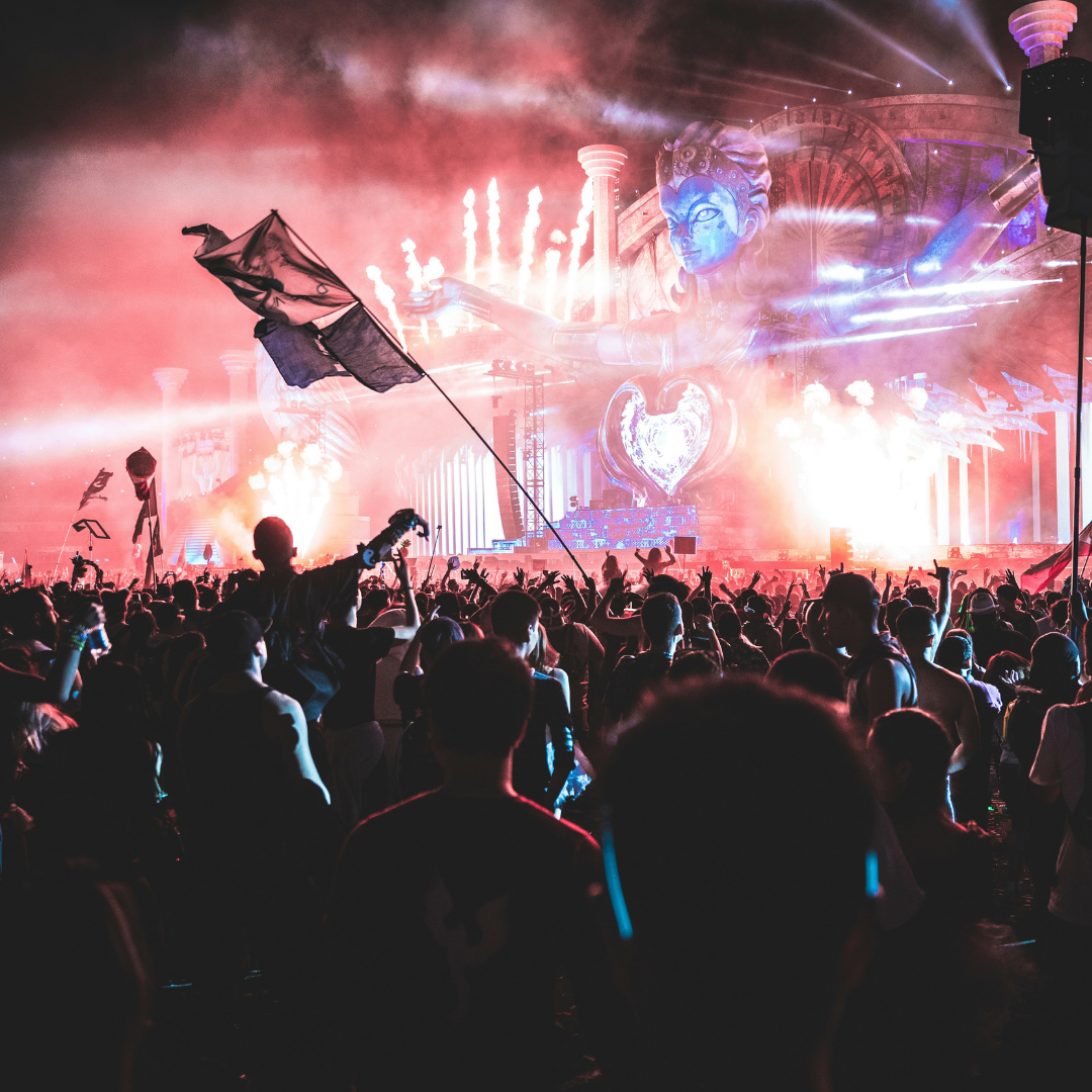 Festival crowd dancing and waving flags in front of a giant illuminated stage statue with pyrotechnics