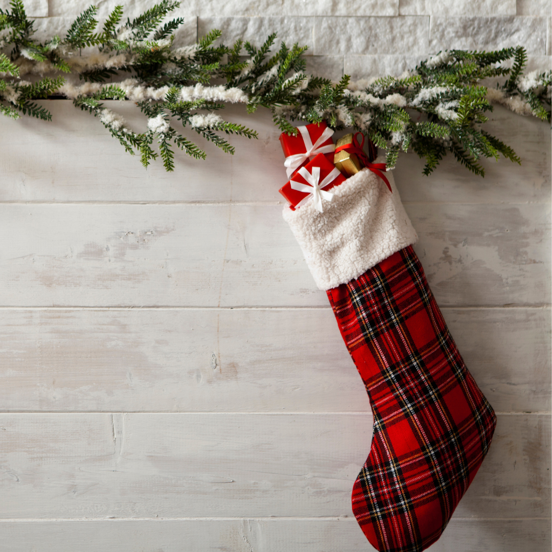 Red plaid Christmas stocking filled with small wrapped gifts hanging beside snow-dusted pine garland on a whitewashed wood background