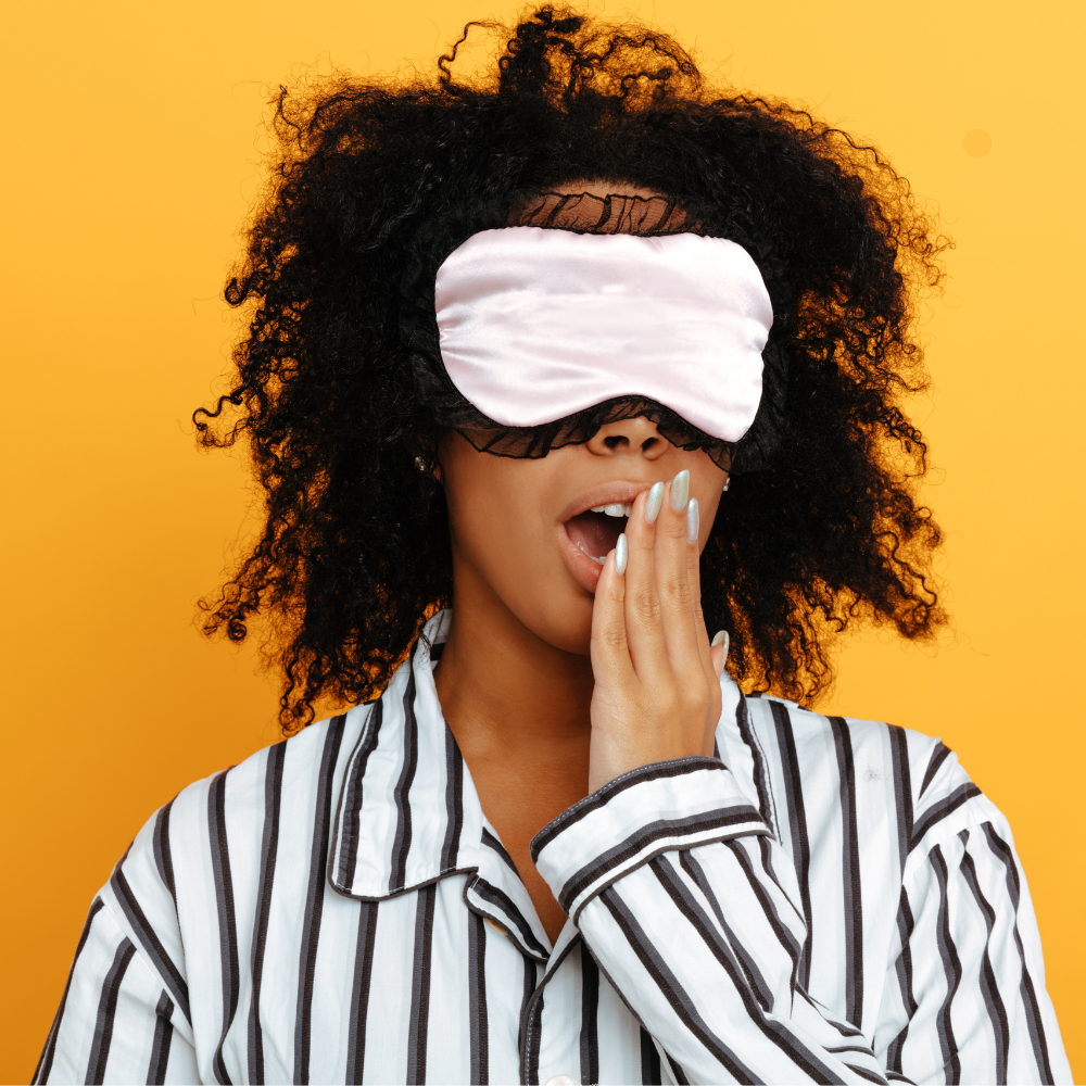 Woman in striped pajamas yawning with hand over her mouth while wearing a pink silk sleep mask against a yellow background