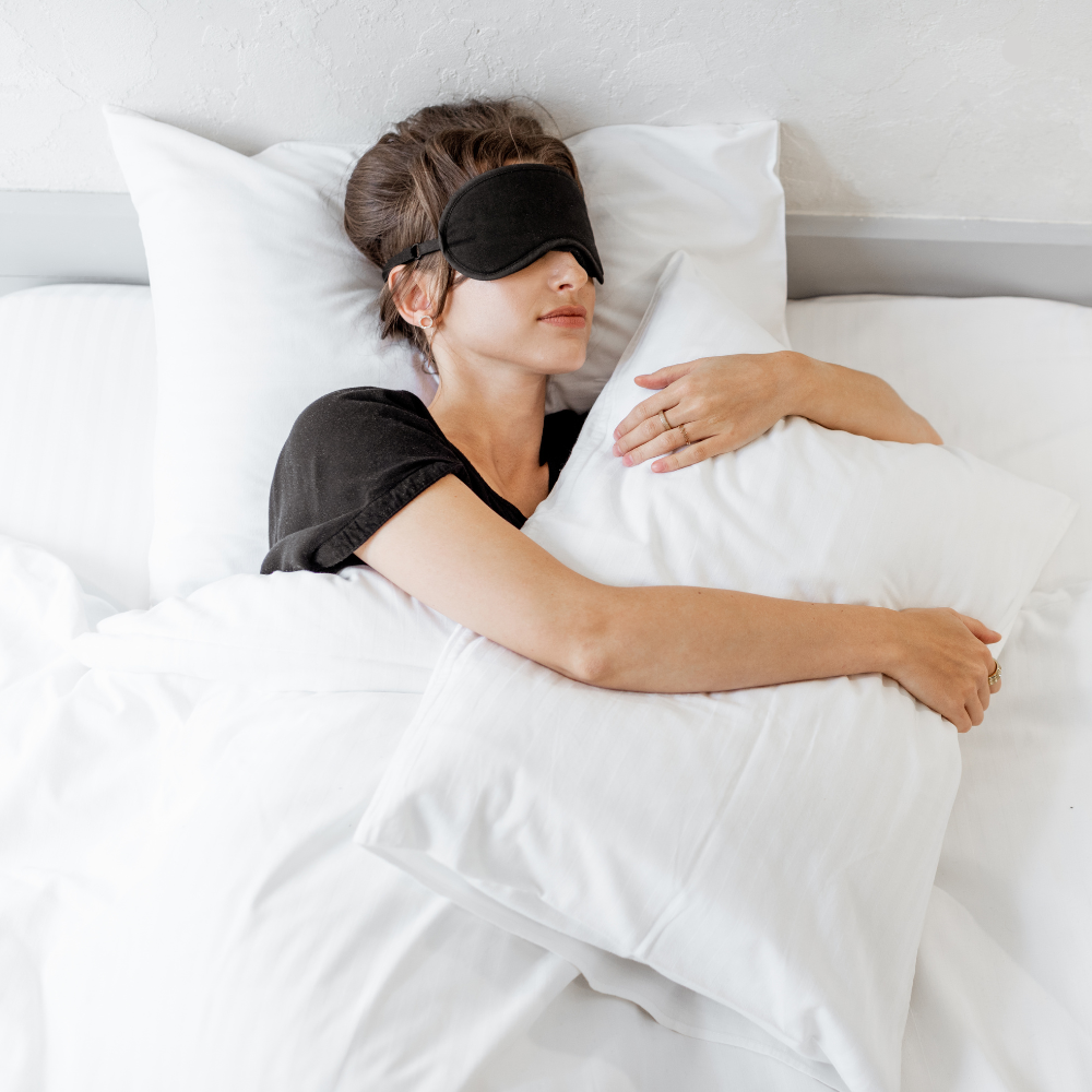 Woman sleeping with a black eye mask hugging a white pillow in a bright bed, illustrating restful sleep and breathing