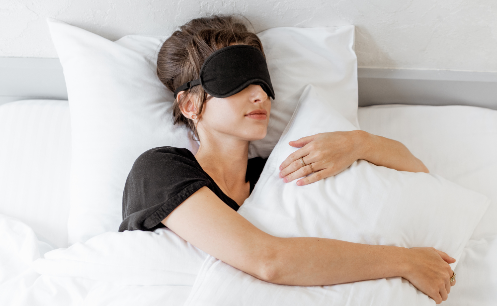 Woman sleeping with a black eye mask, hugging a pillow in a white bed