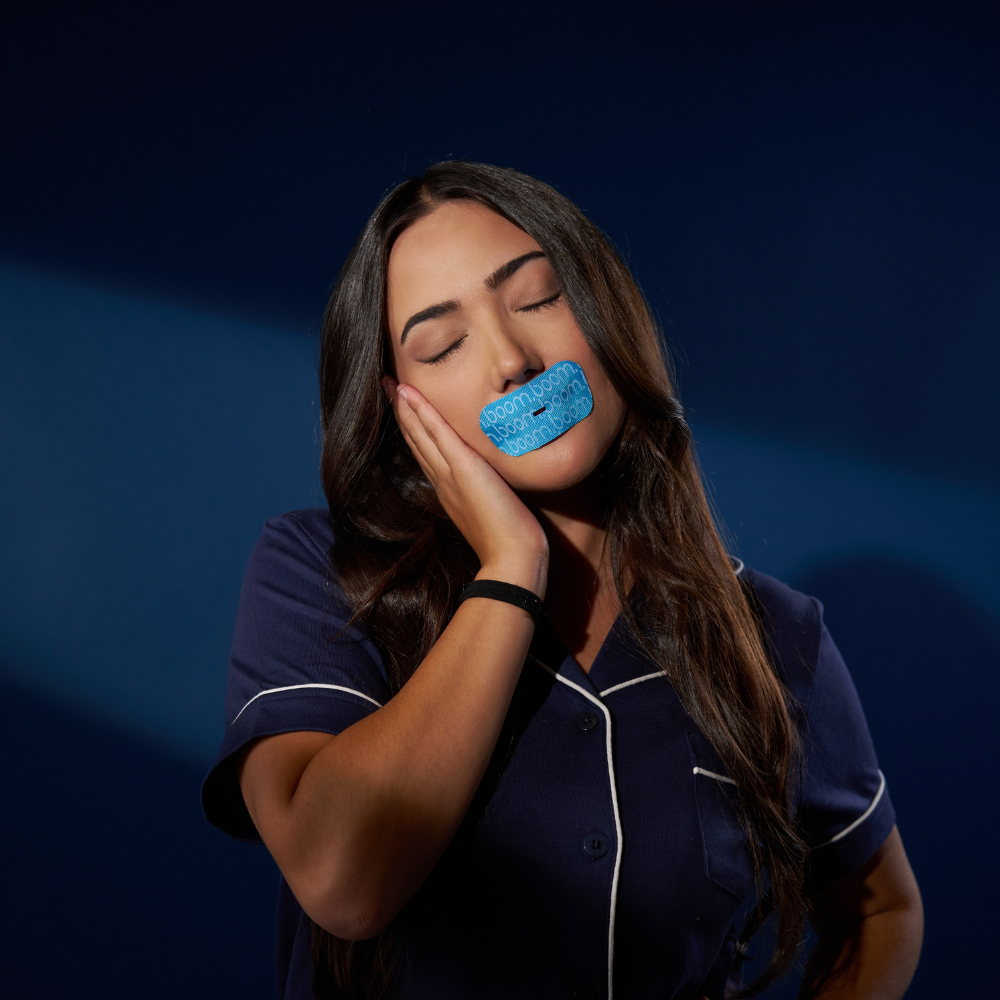 Young woman with eyes closed wearing blue mouth tape, resting her cheek on her hand in navy pajamas against a dark blue background